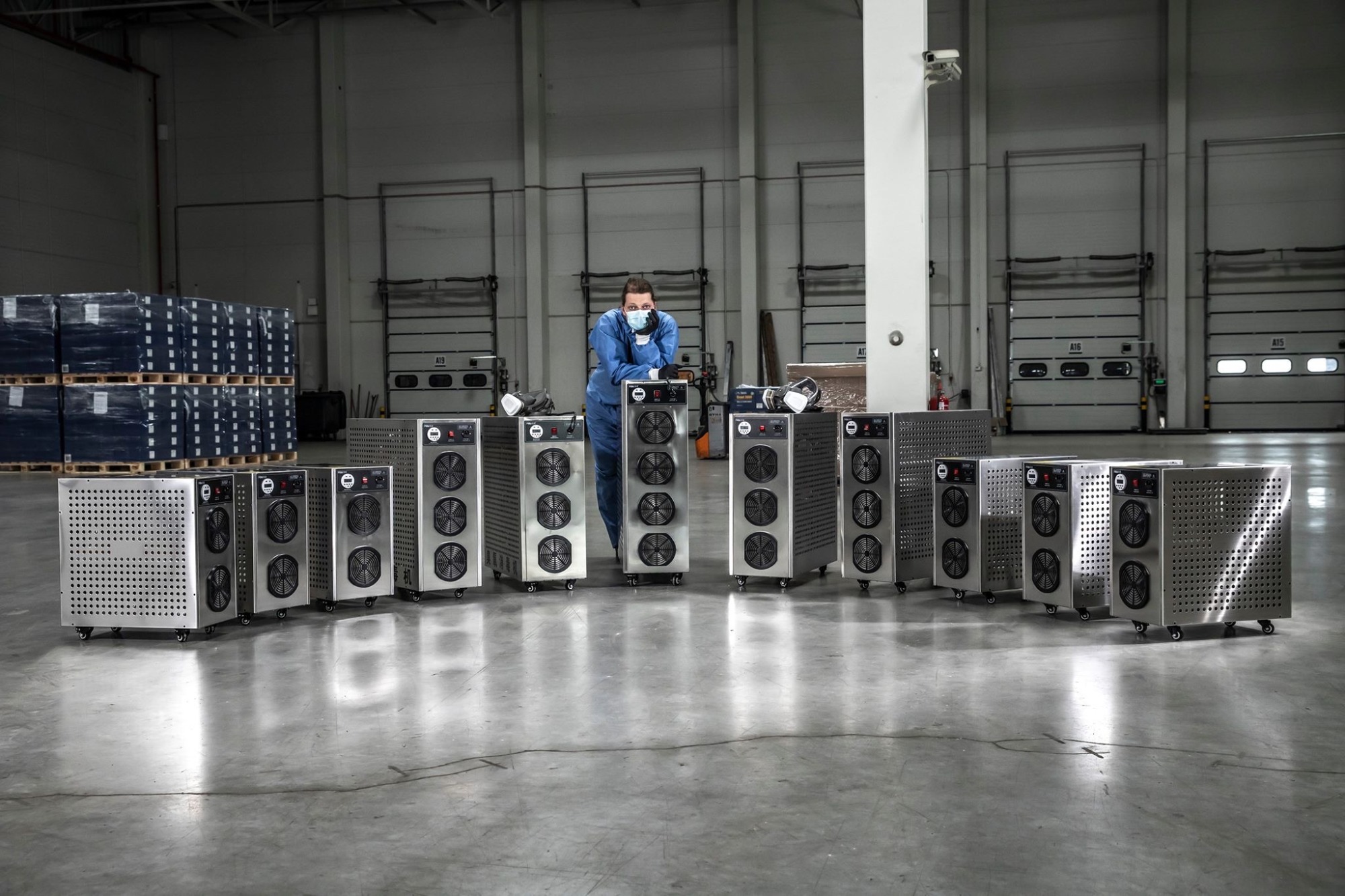 Pracownik w kombinezonie i masce opiera się o mobilne urządzenie z wentylatorami, ustawione w rzędzie na tle magazynu z bramami i paletami.