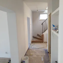 Korytarz w trakcie malowania na biało, zabezpieczona podłoga, widoczny kubeł z farbą i narzędzia malarskie, schody w tle.