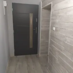 Wnętrze przedpokoju z ciemnymi drzwiami wejściowymi, ścianą wyłożoną panelami imitującymi drewno oraz podłogą z paneli w odcieniach szarości.