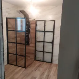 Nowoczesna kabina prysznicowa typu walk-in z czarnymi profilami i deszczownicą, wykończona płytkami imitującymi marmur i drewno w trakcie remontu łazienki.
