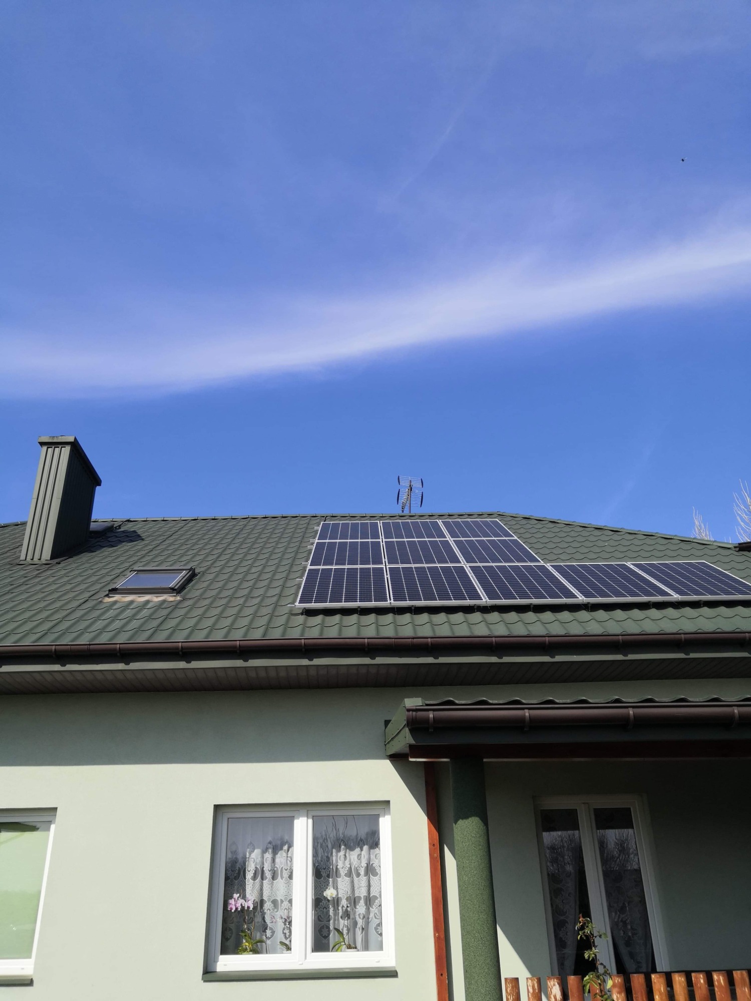 Panele słoneczne na zielonym dachu domu jednorodzinnego z kominem i oknem dachowym, na tle błękitnego nieba z delikatnymi chmurami.