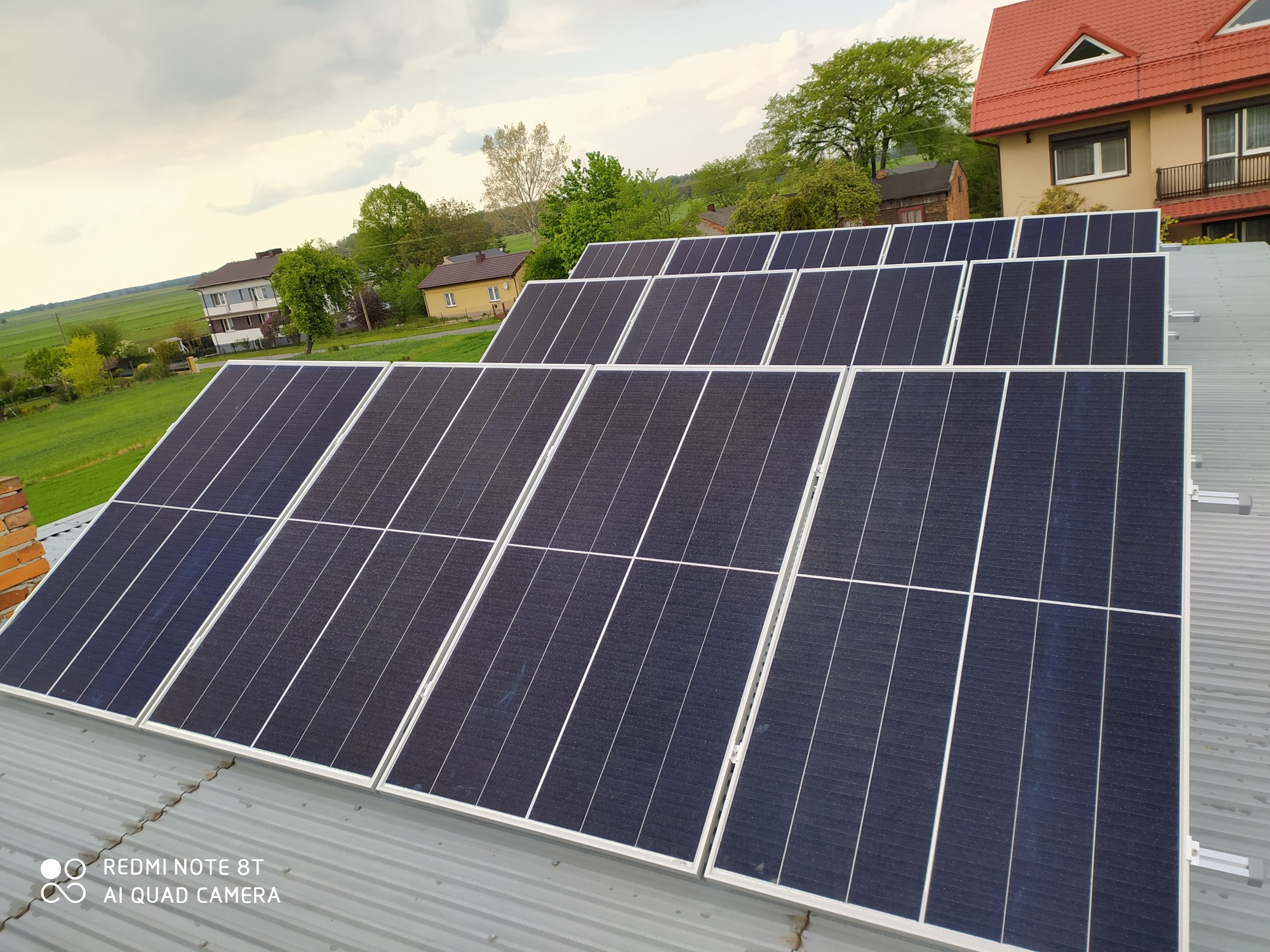 Rząd paneli słonecznych z białymi ramami, zamontowanych na metalowym dachu, z widokiem na wiejski krajobraz i budynki w tle.