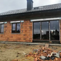 Montaż okien PCV i drzwi przesuwnych w surowym budynku z cegły, widoczne rolokasety, gruz na pierwszym planie.