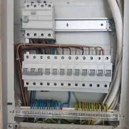 Wnętrze szarej skrzynki rozdzielczej z zainstalowanymi bezpiecznikami Hager, przewodami i zaciskami, widok z bliska na połączenia elektryczne.