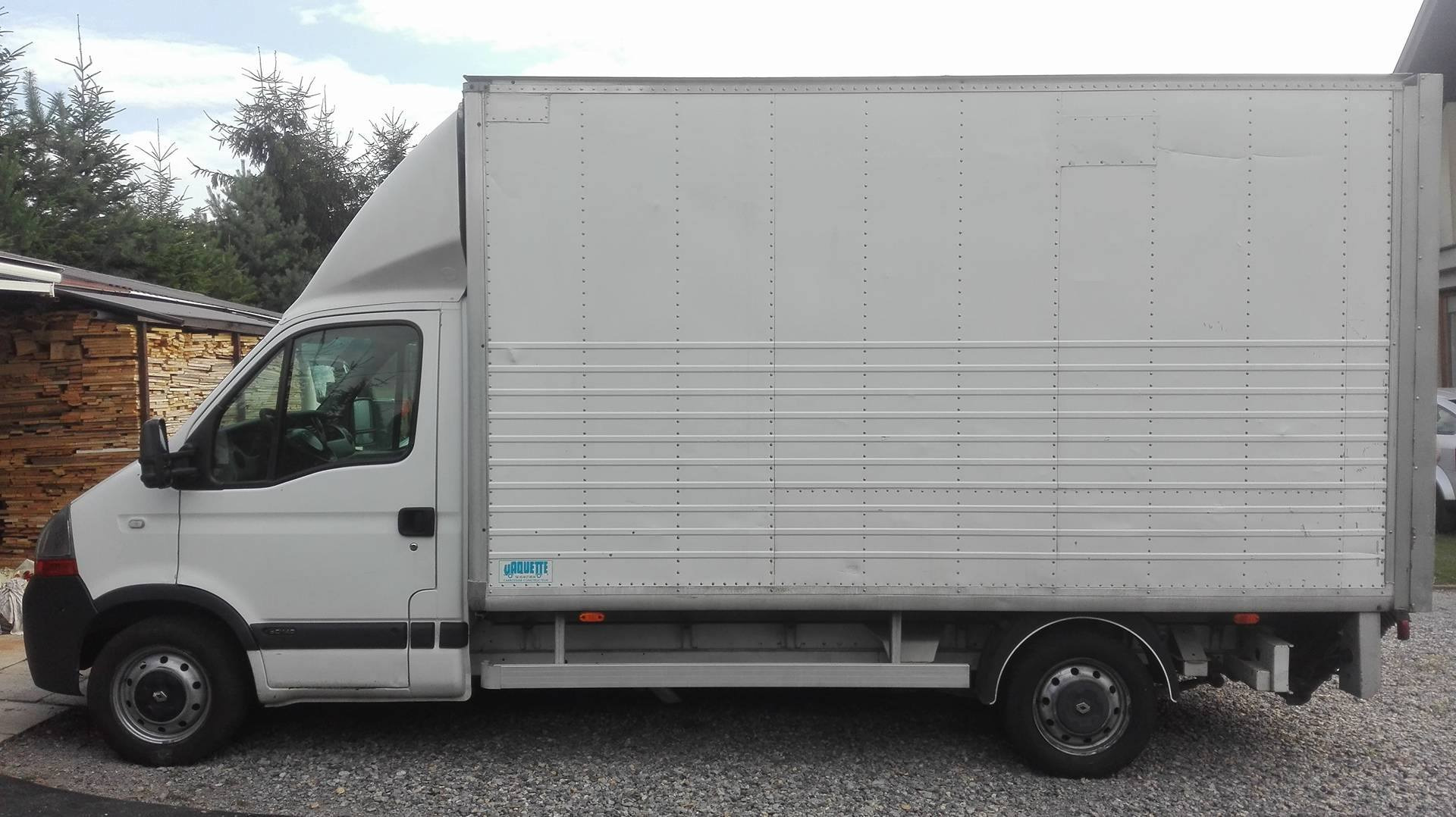 Biały samochód dostawczy z zabudową kontenerową, widok z boku na tle drzew i drewnianej konstrukcji.