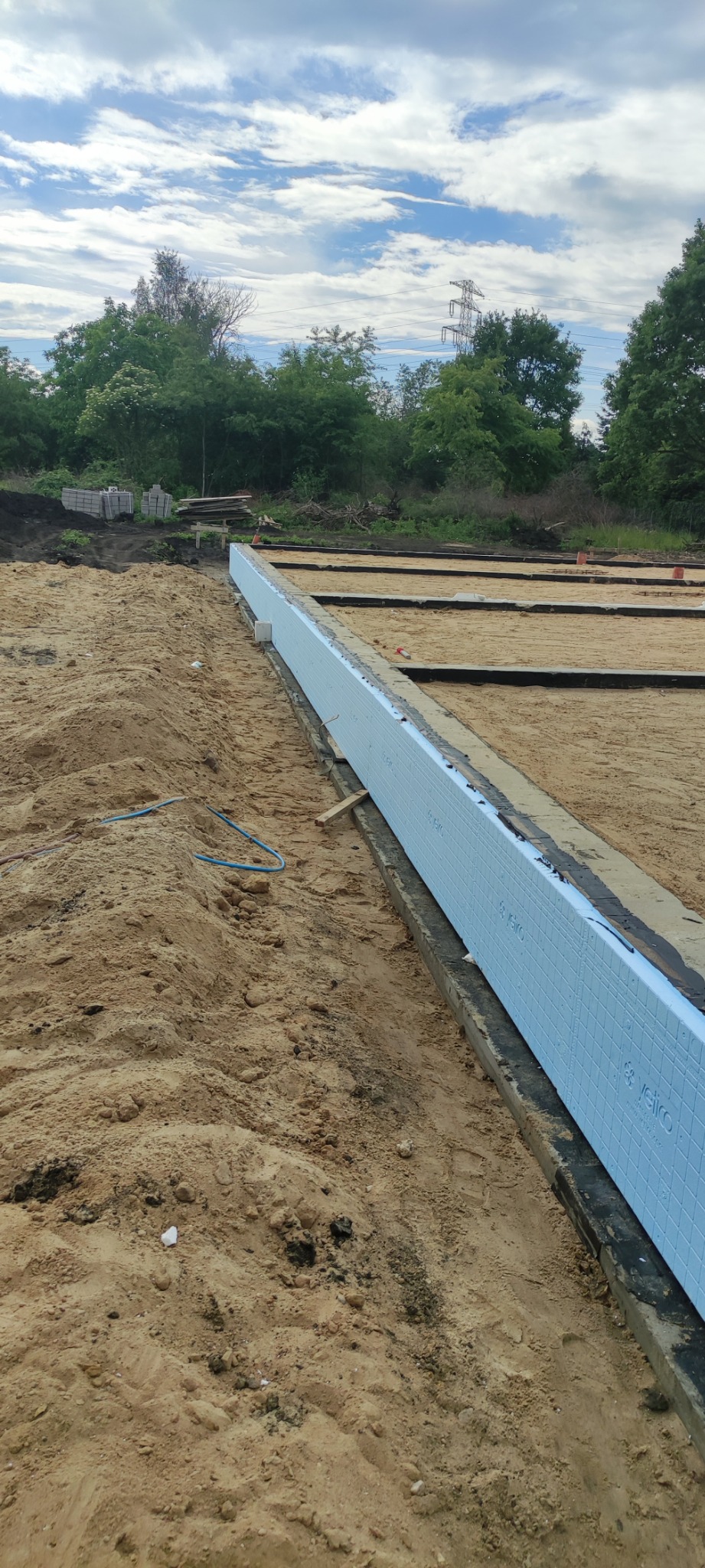 Przygotowanie do wylewki fundamentów: widoczne szalunki i izolacja termiczna z polistyrenu ekstrudowanego, piasek, teren budowy z drzewami w tle.