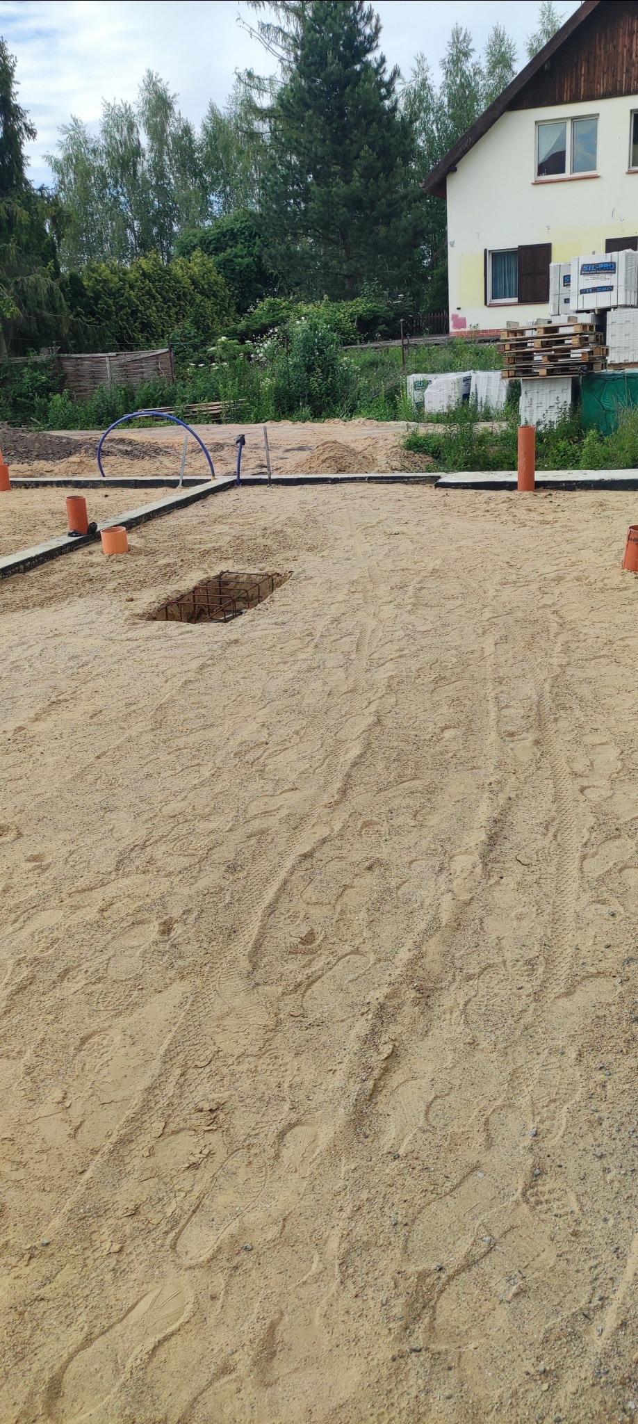 Przygotowanie terenu pod fundamenty: wykop z widocznym zbrojeniem, obrys fundamentów wyznaczony betonowymi obrzeżami i pomarańczowymi rurami, w tle dom jednorodzinny i drzewa.