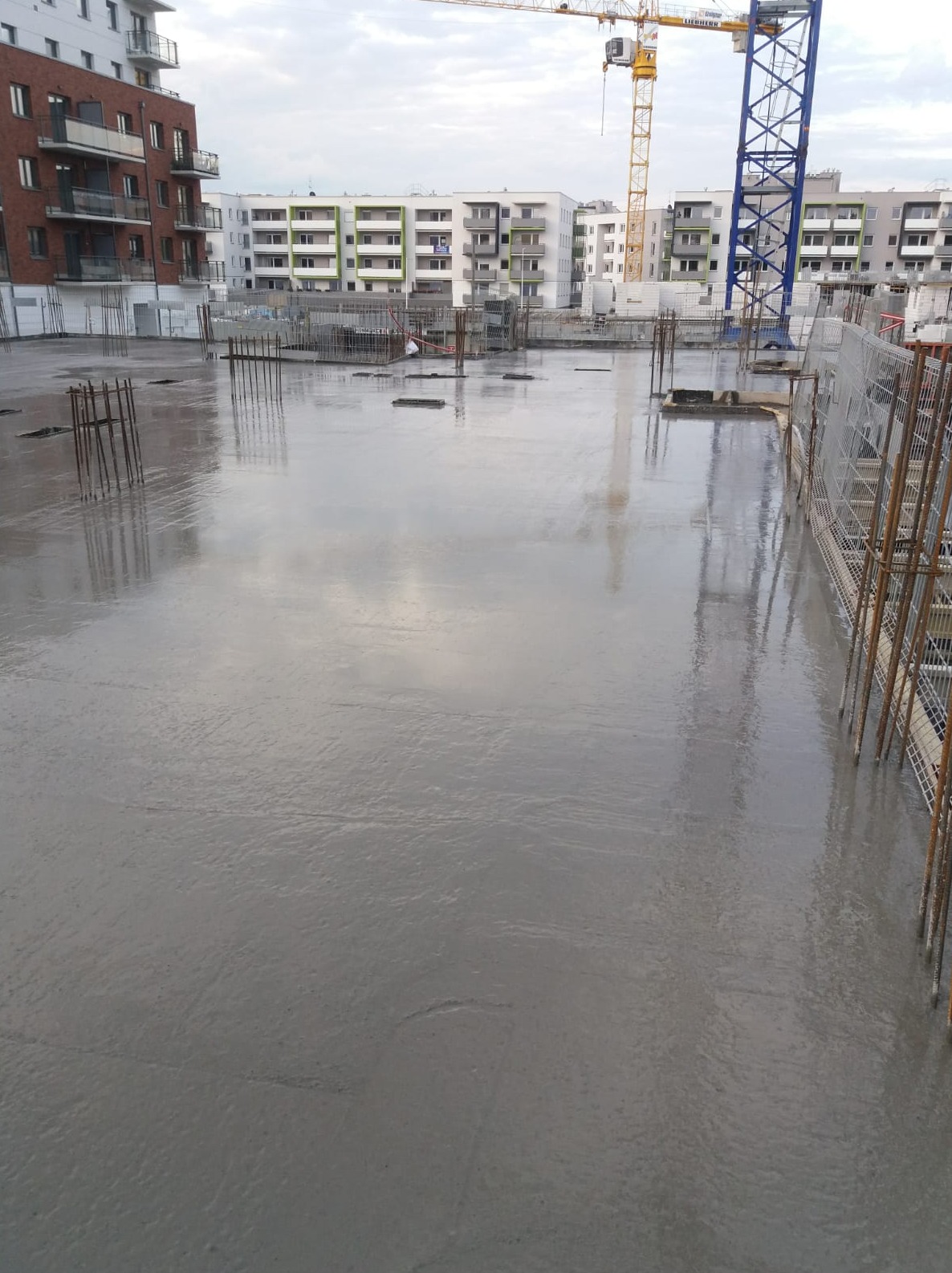 Świeżo wylany, gładki betonowy fundament na budowie z widocznymi wystającymi prętami zbrojeniowymi i żurawiem w tle.