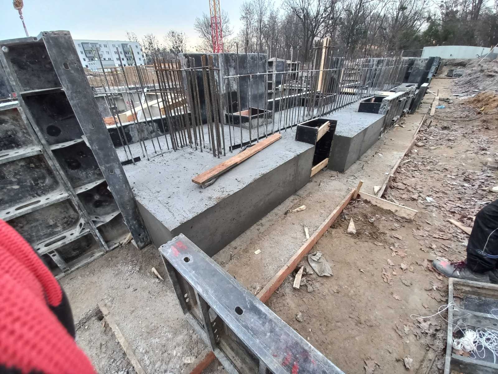Wylane fundamenty z szalunkami i wystającymi prętami zbrojeniowymi na placu budowy, widoczne elementy konstrukcyjne i fragment krajobrazu miejskiego w tle.