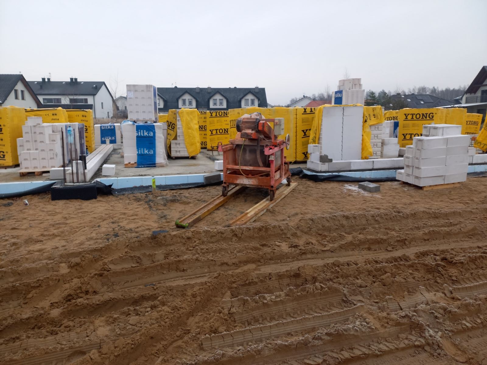 Przygotowanie do budowy fundamentów: widoczny wykop, bloczki fundamentowe Silka i Ytong na paletach, oraz betoniarka na placu budowy.