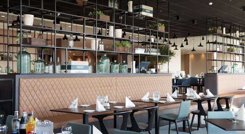 Wnętrze restauracji z rzędem stolików, tapicerowaną ławą pod ścianą i metalowym regałem wiszącym nad nią, wypełnionym dekoracyjnymi naczyniami i roślinami.