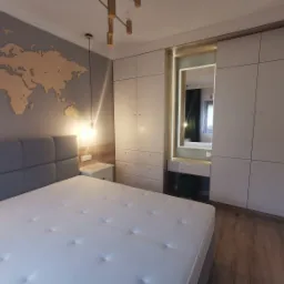 Sypialnia z szarą tapetą z mapą świata, łóżkiem z szarym zagłówkiem, białą szafą z lustrem i toaletką oraz lampą sufitową z elementami w kolorze złota.