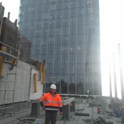 Pracownik w kasku i odblaskowej kurtce na dachu wieżowca podczas układania papy, w tle szklana fasada budynku i dźwig budowlany.