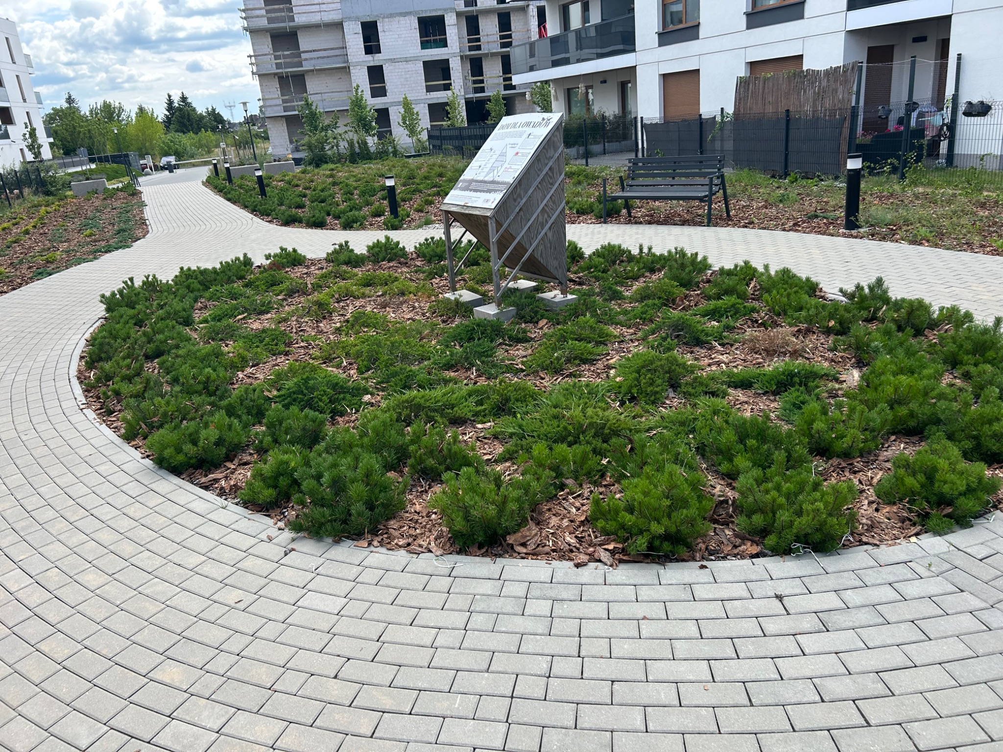 Okrągły klomb z iglakami i korą otoczony brukowaną ścieżką w nowym osiedlu, z tablicą informacyjną i ławką w tle, w słoneczny dzień.