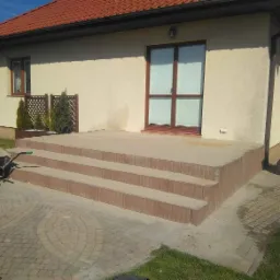 Betonowy taras z trzema stopniami z kostki, przylegający do budynku z beżową elewacją i brązowymi oknami, widoczny wózek budowlany i narzędzia, kostka brukowa na podjeździe.