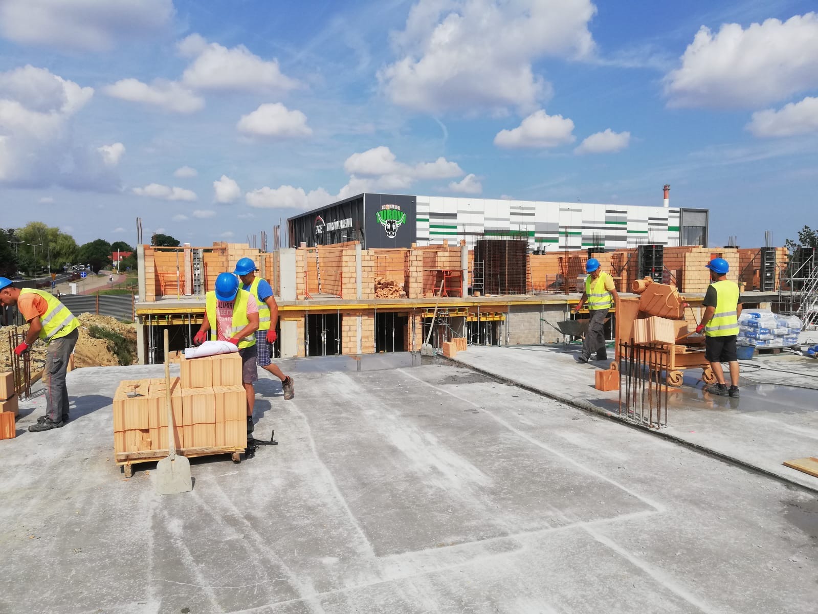 Pracownicy budowlani w kaskach i kamizelkach odblaskowych układają cegły na budowie, w tle widoczny budynek z logo.