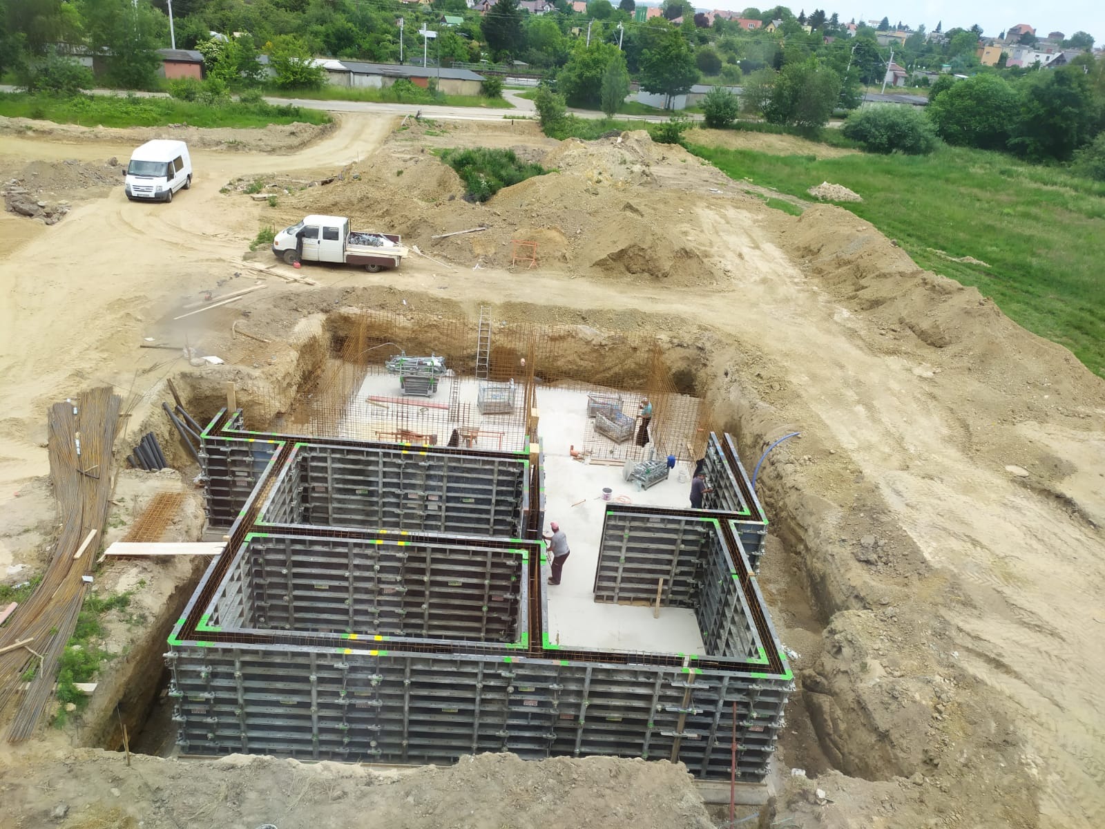 Budowa fundamentów domu jednorodzinnego: szalunki zbrojeniowe w wykopie, widoczne stalowe pręty i betonowa podstawa, na zewnątrz wykopu zaparkowane samochody dostawcze.