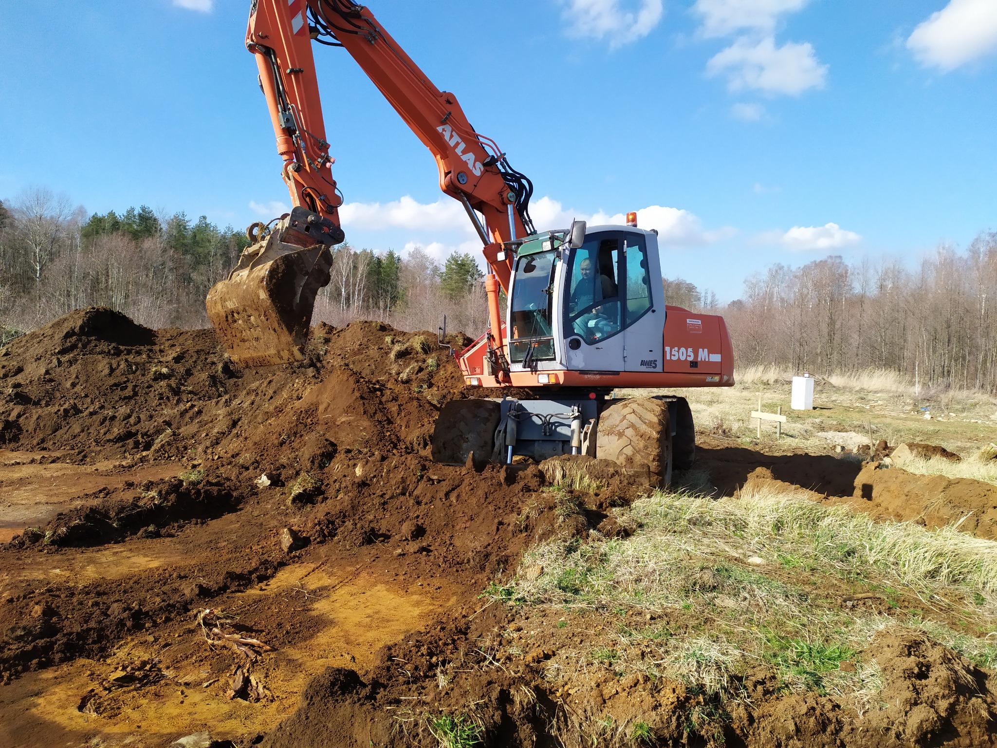 Pomarańczowa koparka Atlas 1505 M nabiera ziemię z wykopu na tle lasu i błękitnego nieba z chmurami.