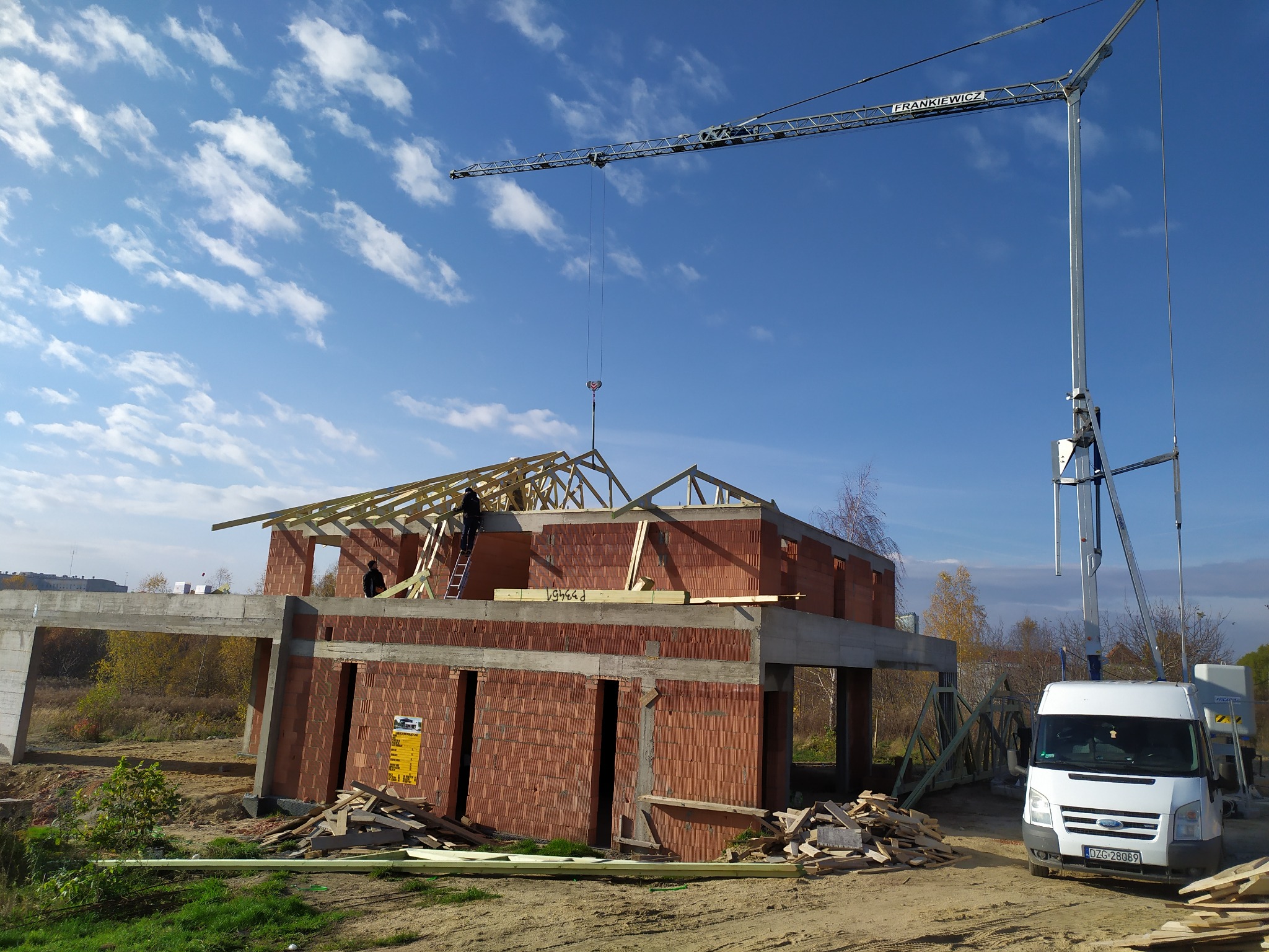 Budowa domu jednorodzinnego w stanie surowym otwartym: mur z czerwonej cegły, drewniana konstrukcja dachu w trakcie montażu, dźwig budowlany na tle błękitnego nieba.