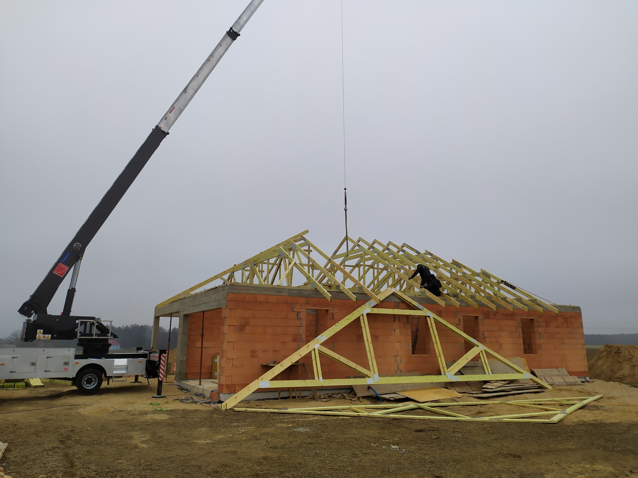 Montaż drewnianej konstrukcji dachu domu jednorodzinnego z użyciem dźwigu; widoczny mur z cegieł i dwie osoby pracujące na wysokości.