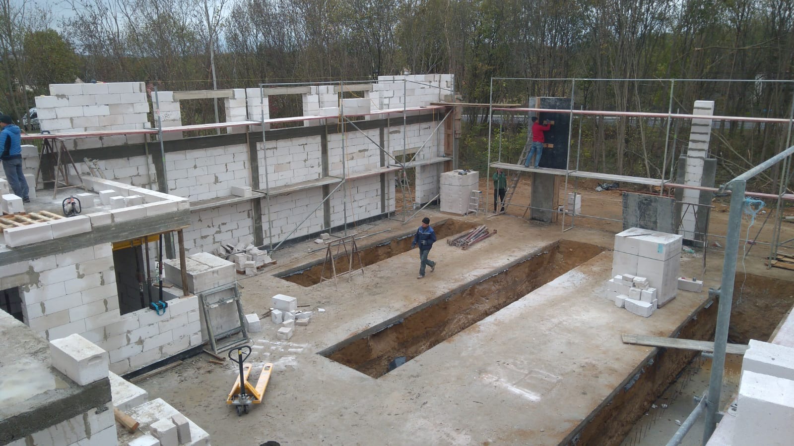Budowa domu jednorodzinnego w trakcie wznoszenia ścian z bloczków betonowych; widoczne fundamenty, rusztowania i pracownicy.