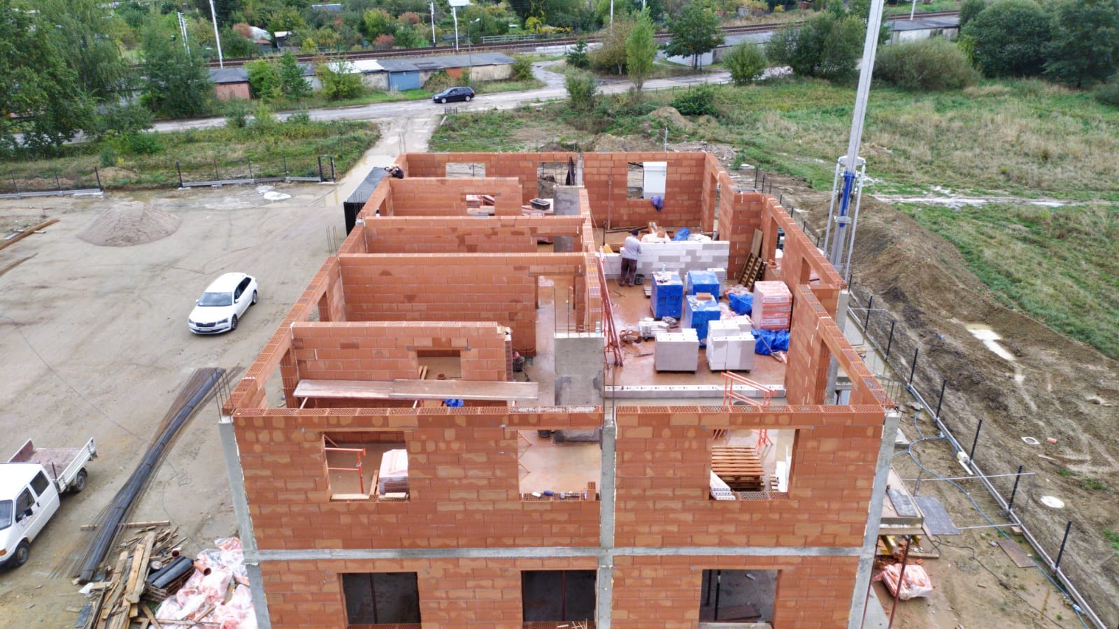 Wznoszenie dwukondygnacyjnego budynku z czerwonej cegły, widok z góry na plac budowy z zaparkowanymi samochodami i stertą materiałów budowlanych.
