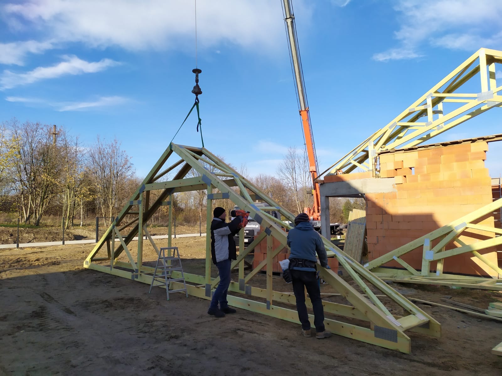 Montaż drewnianej konstrukcji więźby dachowej za pomocą dźwigu; pracownicy mocują elementy, w tle mur z cegły i błękitne niebo.