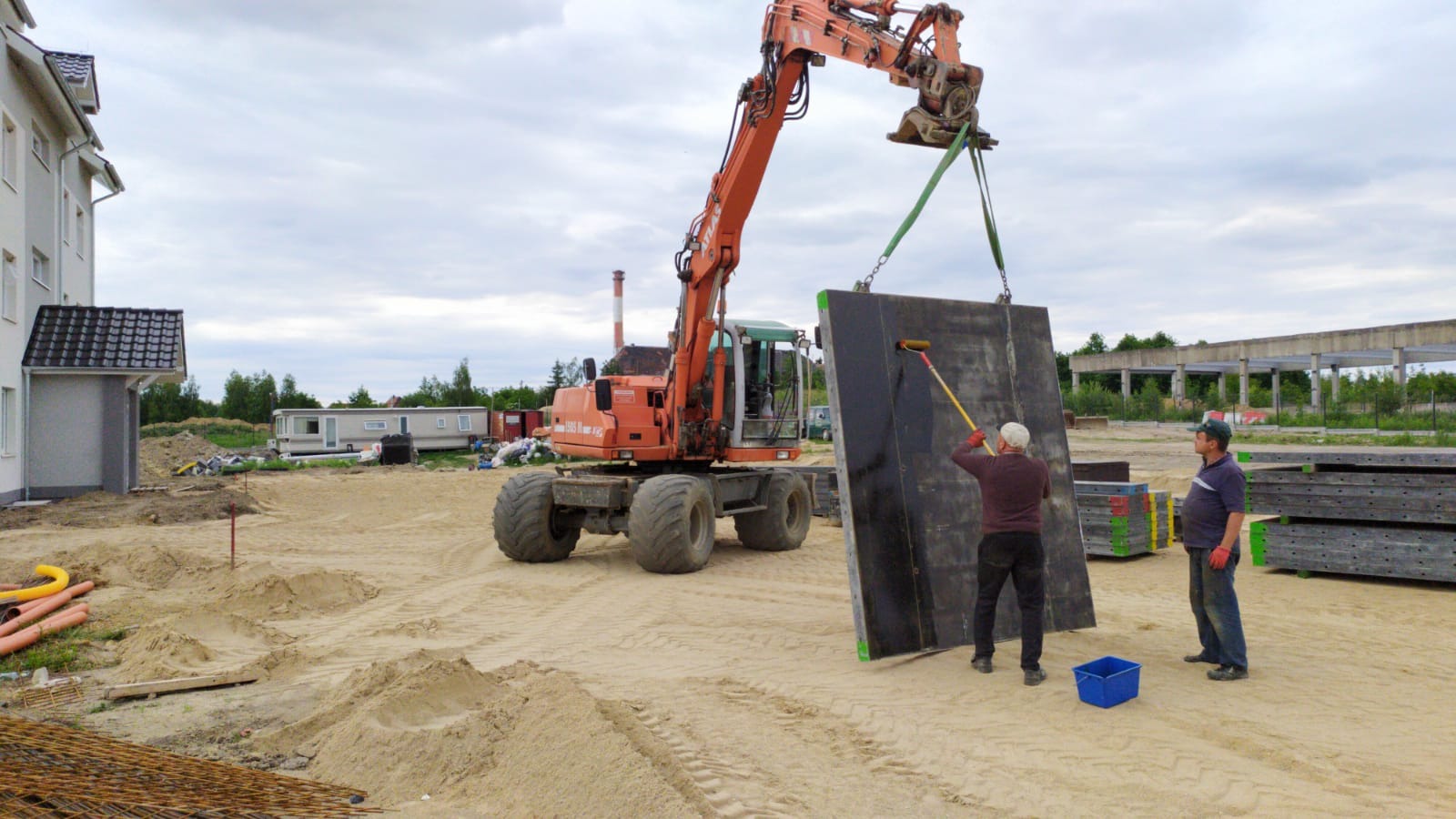 Pomarańczowa koparka podnosi pionowo czarną płytę, którą robotnik maluje wałkiem na budowie, drugi robotnik obserwuje.
