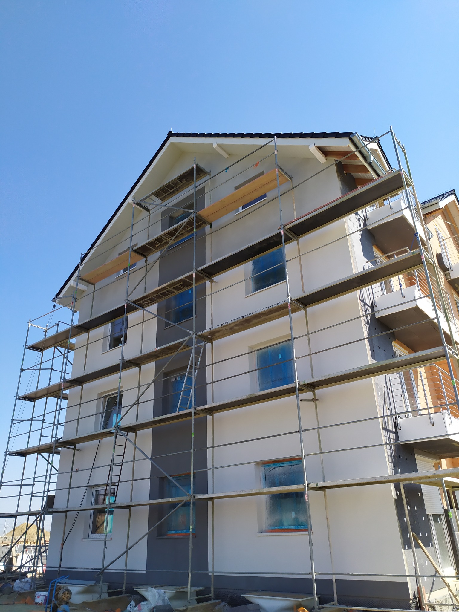 Budynek wielorodzinny w trakcie ocieplania elewacji, zabezpieczone okna folią, rusztowania na całej wysokości, widoczny fragment dachu z rynną.