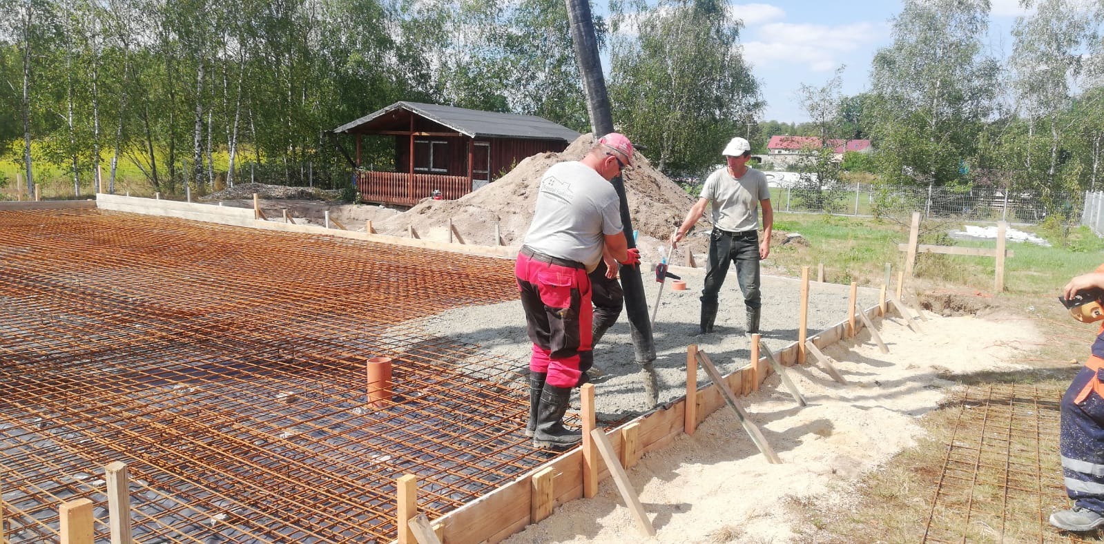 Wylewanie betonu na przygotowane zbrojenie pod fundament budynku jednorodzinnego, widoczna drewniana szalówka i pracownicy w roboczych ubraniach, w tle domek letniskowy.
