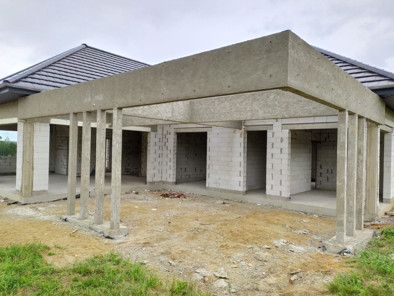 Surowy stan budynku z szarej dachówki, betonowymi słupami i ścianami z białych bloczków, widoczne z zewnątrz na tle pochmurnego nieba.