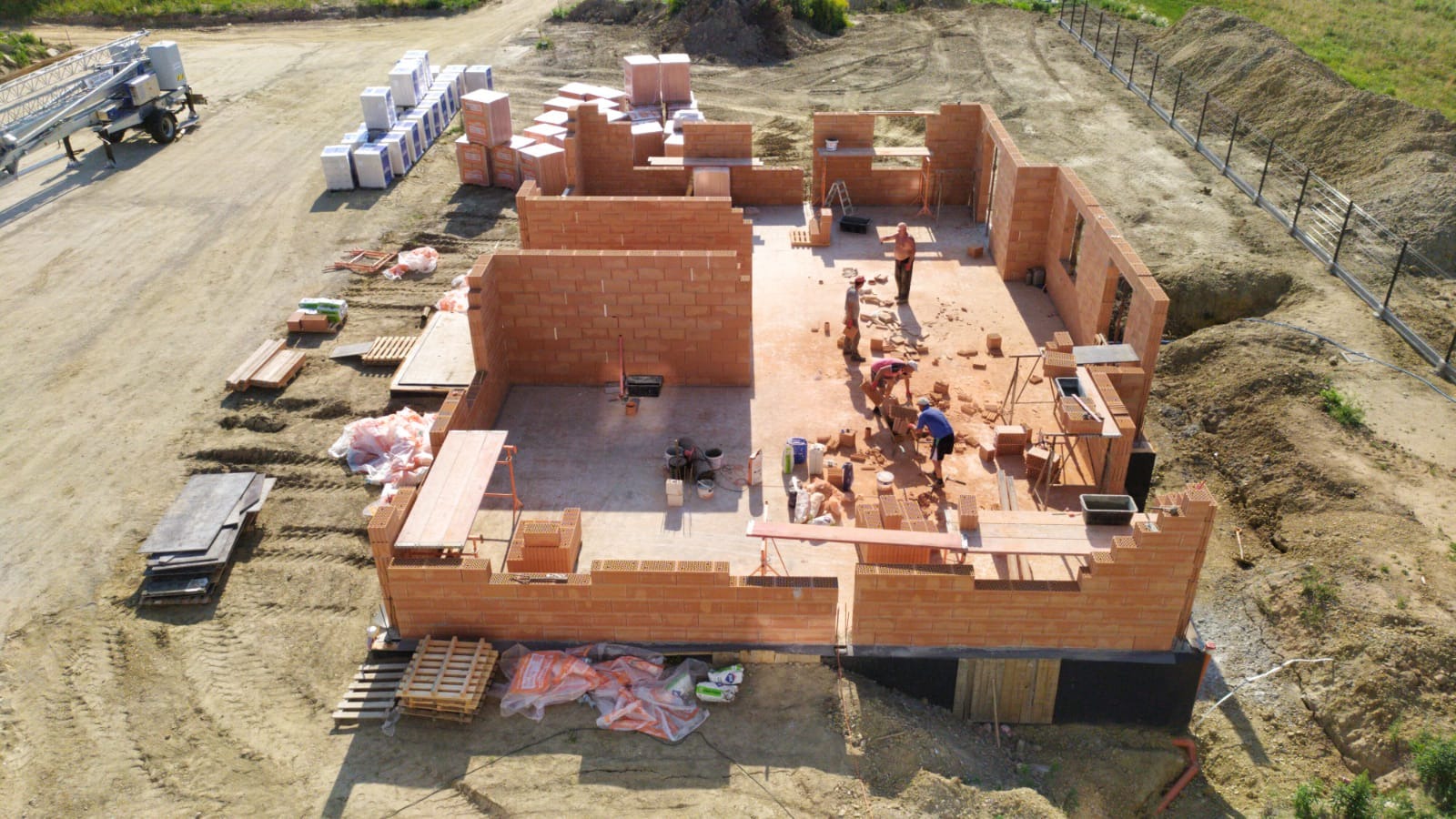 Budowa domu jednorodzinnego z czerwonej cegły, widok z góry na plac budowy, gdzie murarze pracują nad wznoszeniem ścian, a obok leżą materiały budowlane i drabina na przyczepie.