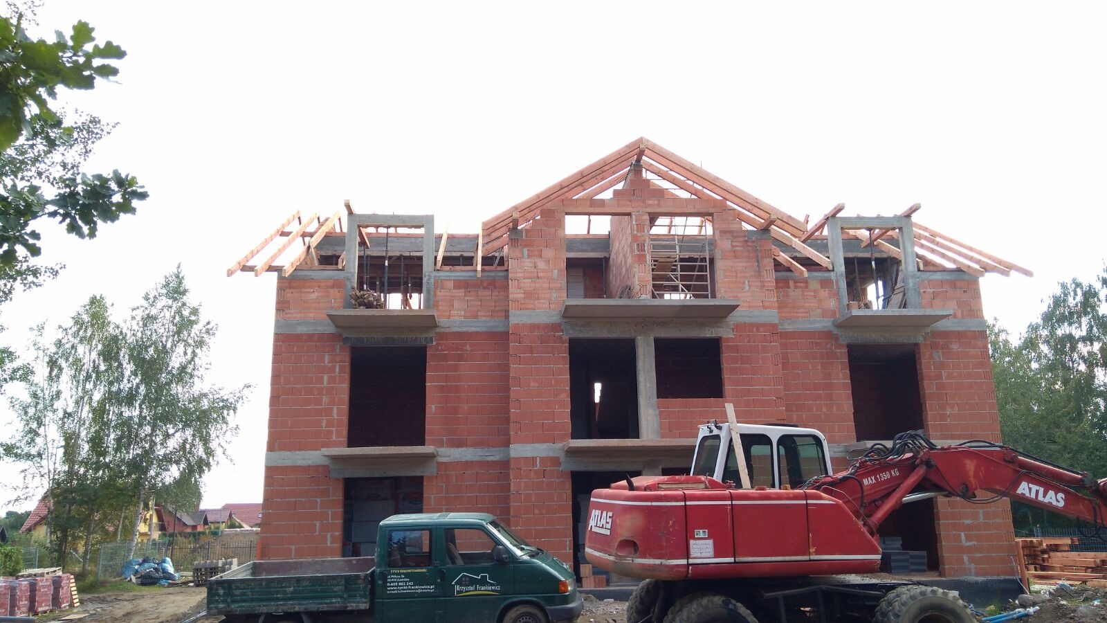 Wznoszenie dwupiętrowego domu z czerwonej cegły, widoczna drewniana konstrukcja dachu, na pierwszym planie czerwona koparka Atlas i zielony samochód dostawczy.