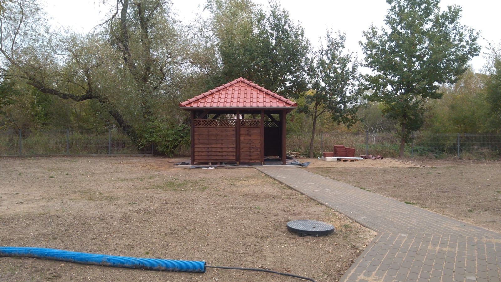 Altana z czerwonym dachem z dachówki ceramicznej, z drewnianymi ściankami z kratką, widok z przodu na ścieżkę z kostki brukowej, w tle drzewa i ogrodzenie z siatki, na ziemi leży niebieski wąż...