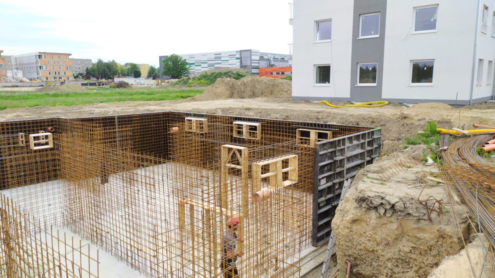 Budowa fundamentów domu: zbrojenie stalowe, szalunki, widoczny pracownik w kasku, w tle budynki mieszkalne.