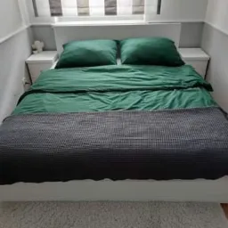 Nowoczesna sypialnia z białym łóżkiem i zagłówkiem, zieloną pościelą i szarą narzutą w wafelkową kratę. Po bokach łóżka białe szafki nocne. U góry okno z żaluzjami.