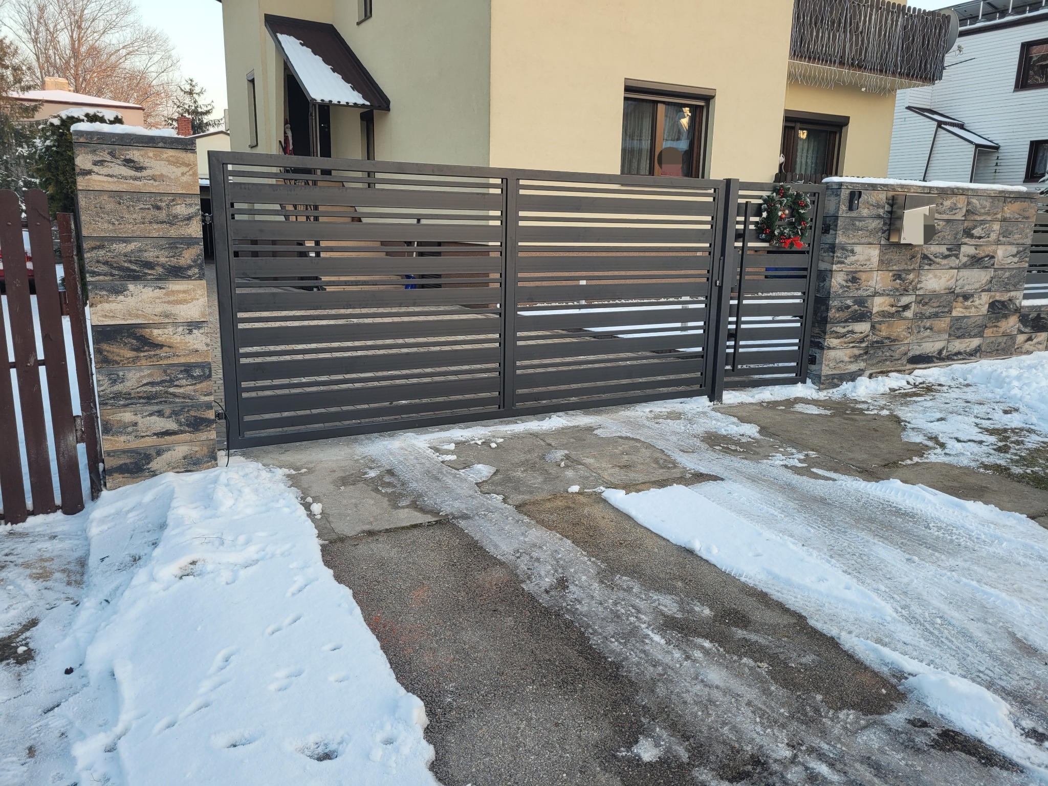 Nowoczesna, pozioma brama wjazdowa z metalowymi panelami w kolorze antracytowym, zamontowana pomiędzy kamiennymi słupkami ogrodzeniowymi. Na jednym ze słupków wisi wieniec świąteczny. Podjazd...