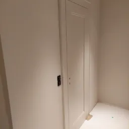 Białe, klasyczne drzwi wewnętrzne z delikatnymi frezowaniami, zamontowane w ścianie o beżowym odcieniu, widoczne elementy montażowe i zabezpieczenia podłogi.