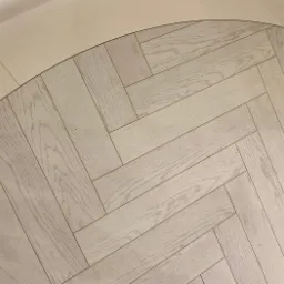 Jasny parkiet ułożony w jodełkę francuską, z widoczną krawędzią wykończeniową w kolorze kremowym.