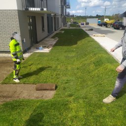 Pit Bud - Układanie trawy z rolki przy nowym budynku mieszkalnym, widoczny pracownik w odblaskowej odzieży i częściowo rozłożona rolka trawy na przygotowanym podłożu.