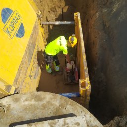 Pit Bud - Pracownik w jaskrawożółtej odzieży roboczej zagęszcza podłoże w głębokim wykopie zabezpieczonym żółtymi szalunkami KOPRAS, widoczna niebieska rura i betonowa płyta z otworami.