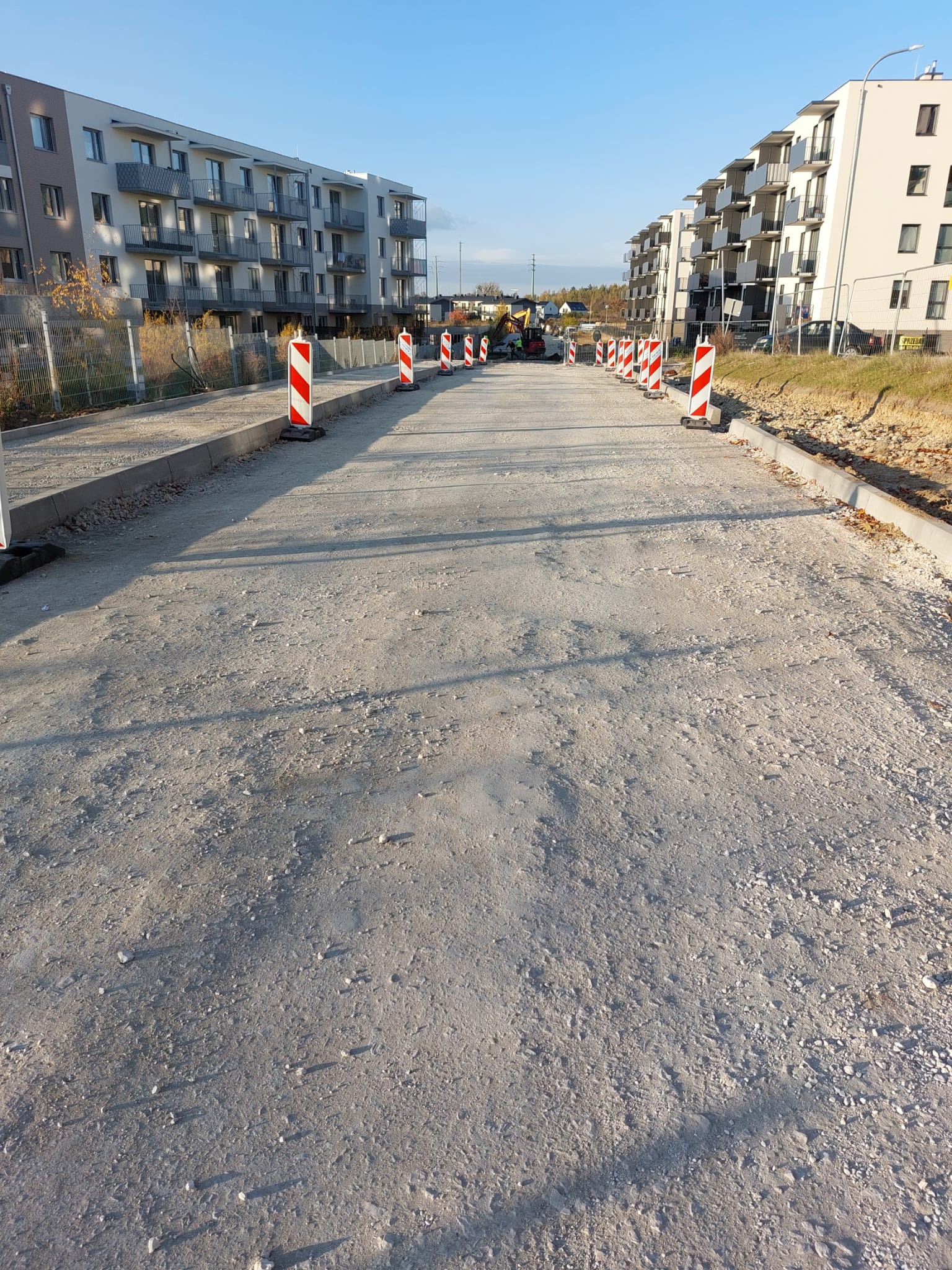 Przygotowany podkład z kruszywa pod przyszłą drogę dojazdową do osiedla mieszkaniowego, widoczne krawężniki i słupki ostrzegawcze.