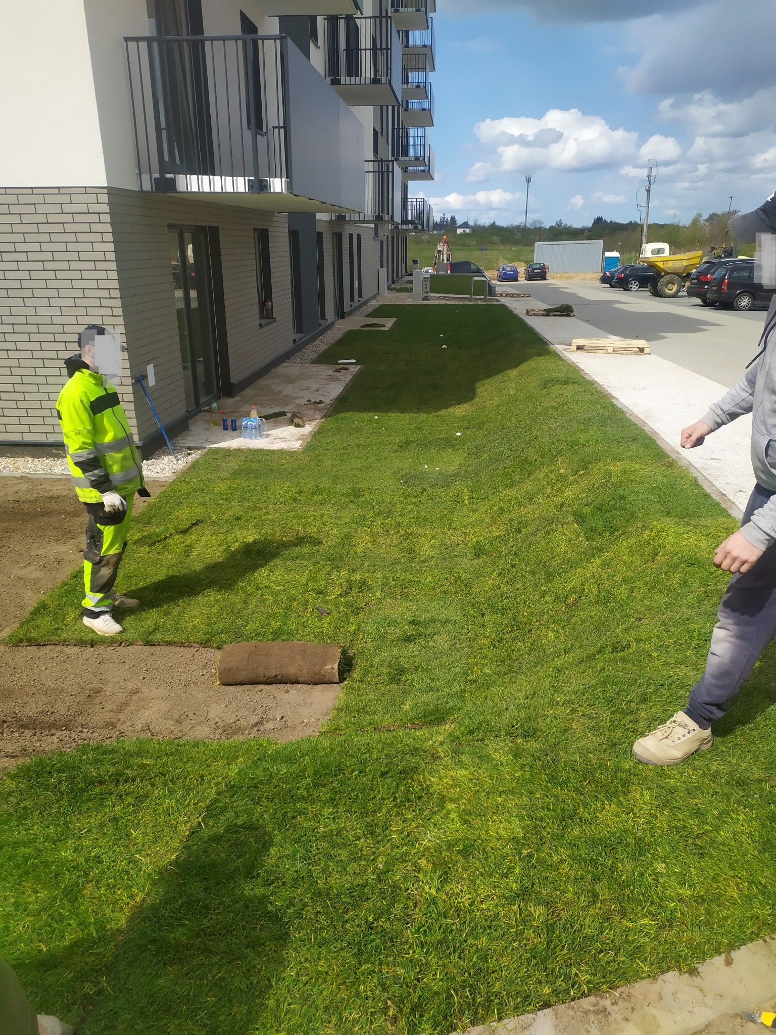 Układanie trawy z rolki przy nowym budynku mieszkalnym, widoczny pracownik w odblaskowej odzieży i częściowo rozłożona rolka trawy na przygotowanym podłożu.