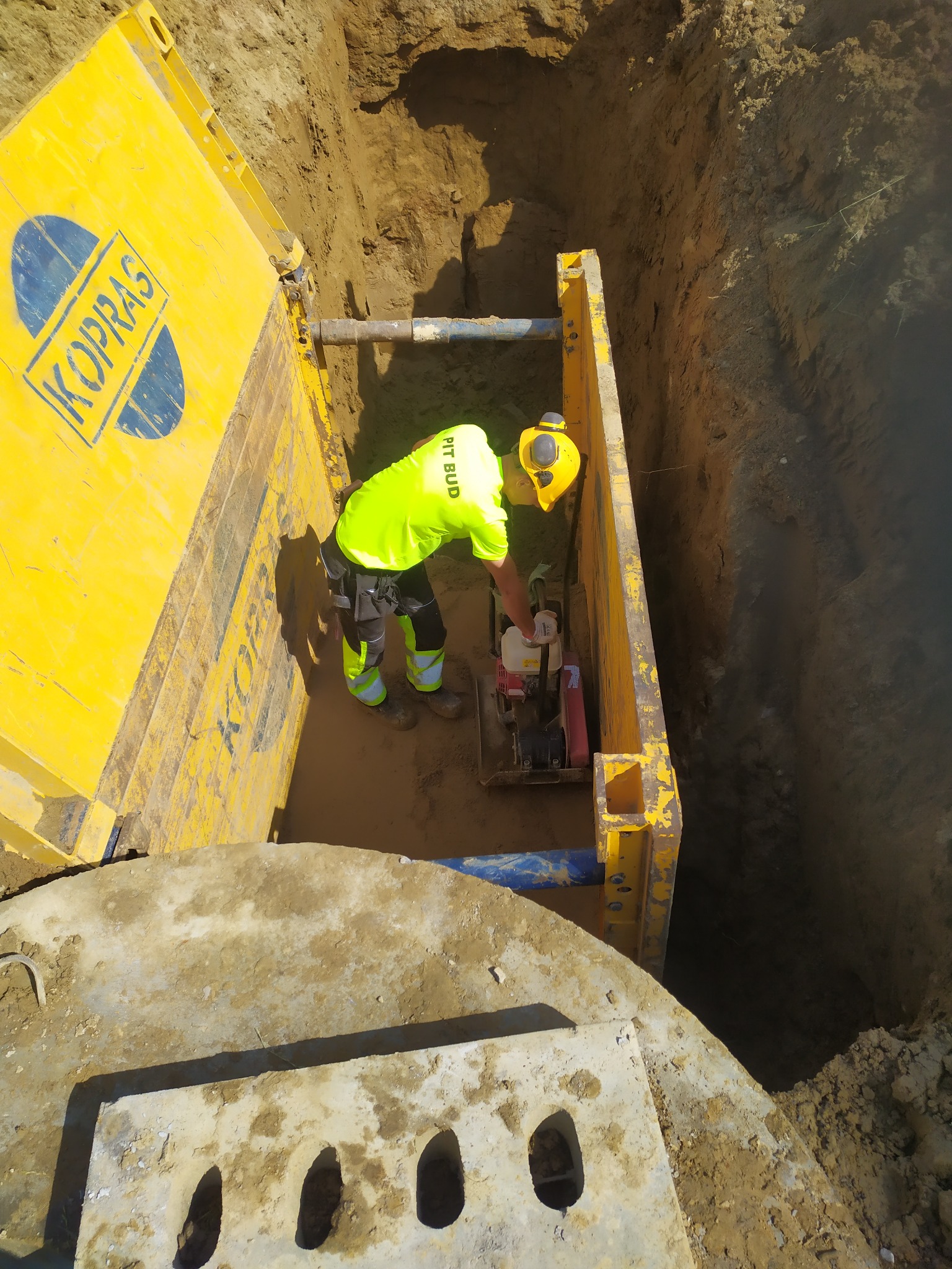 Pracownik w jaskrawożółtej odzieży roboczej zagęszcza podłoże w głębokim wykopie zabezpieczonym żółtymi szalunkami KOPRAS, widoczna niebieska rura i betonowa płyta z otworami.
