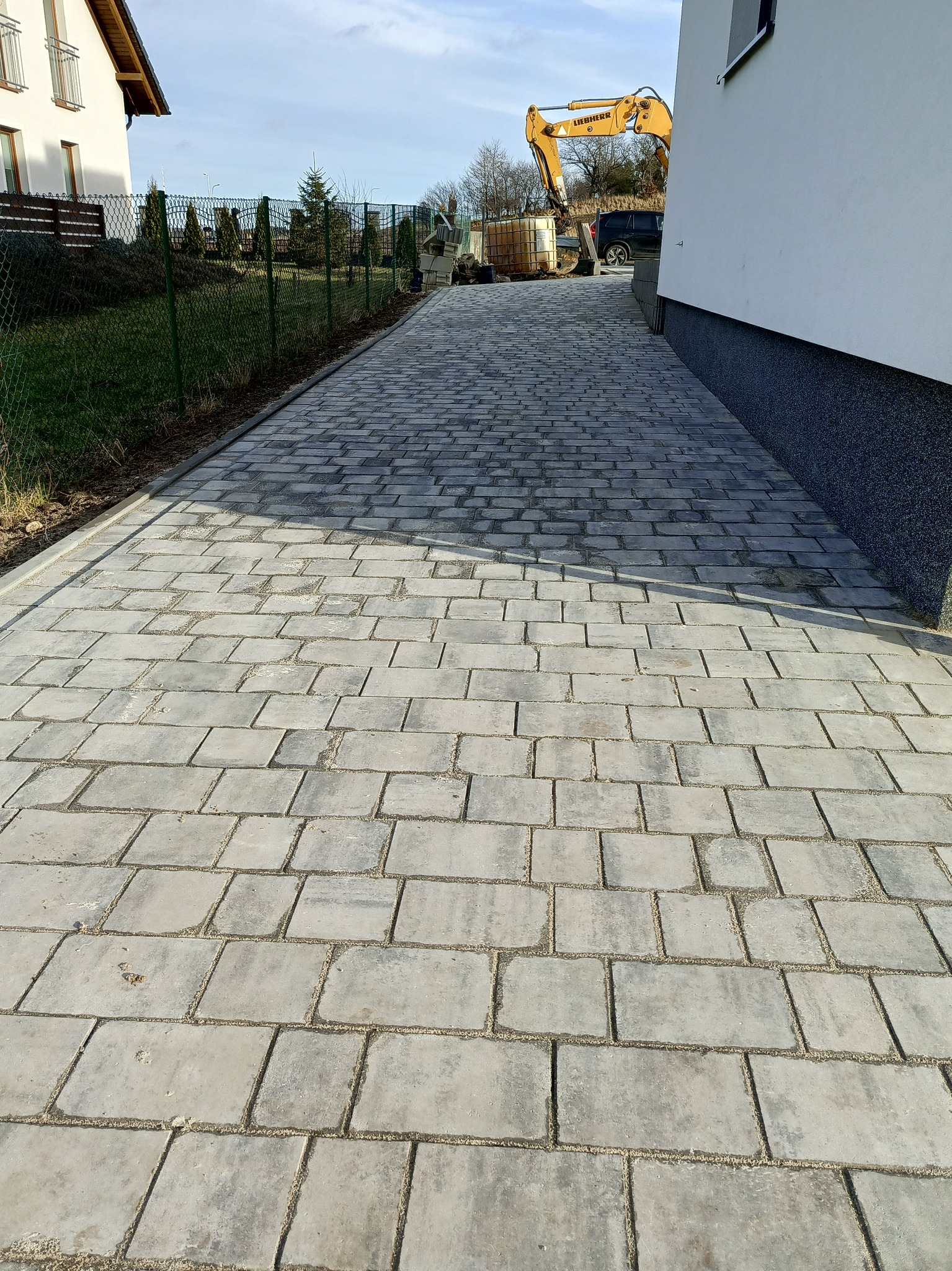 Świeżo ułożona, szara kostka brukowa prowadząca wzdłuż budynku, z widocznym w tle żółtym koparką Liebherr i zielonym ogrodzeniem.