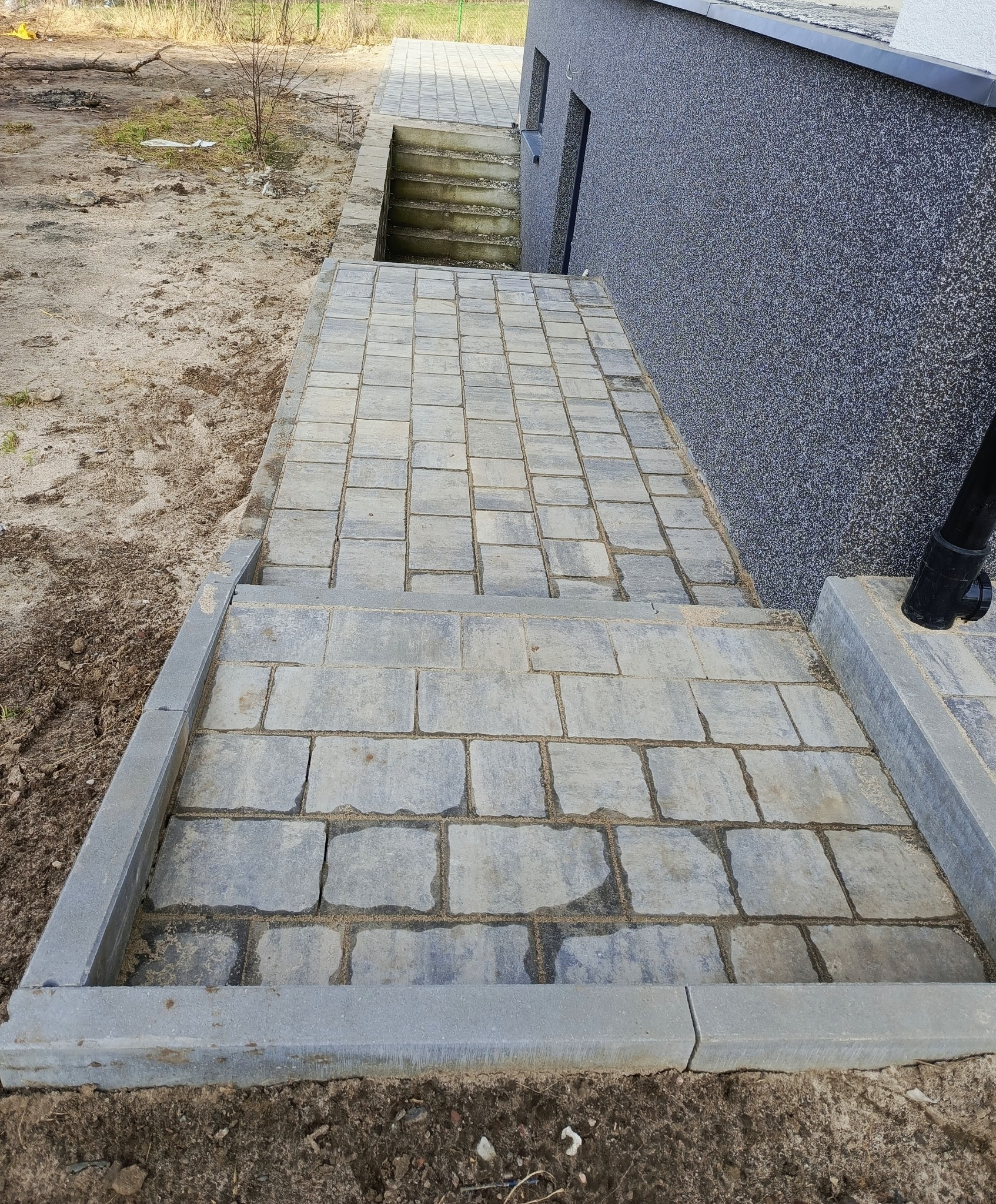 Ułożona ścieżka z szarej kostki brukowej prowadząca do wejścia do budynku, z widocznymi schodami w dół i obrzeżami z kostki.