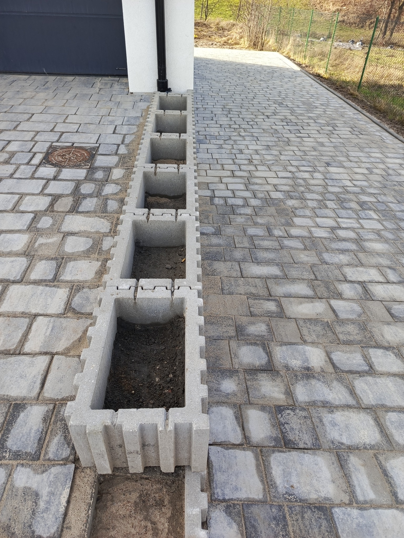 Ułożona kostka brukowa w odcieniach szarości z liniowym systemem drenażowym z betonowych koryt wypełnionych ziemią, widok z góry w kierunku garażu i ogrodzenia.