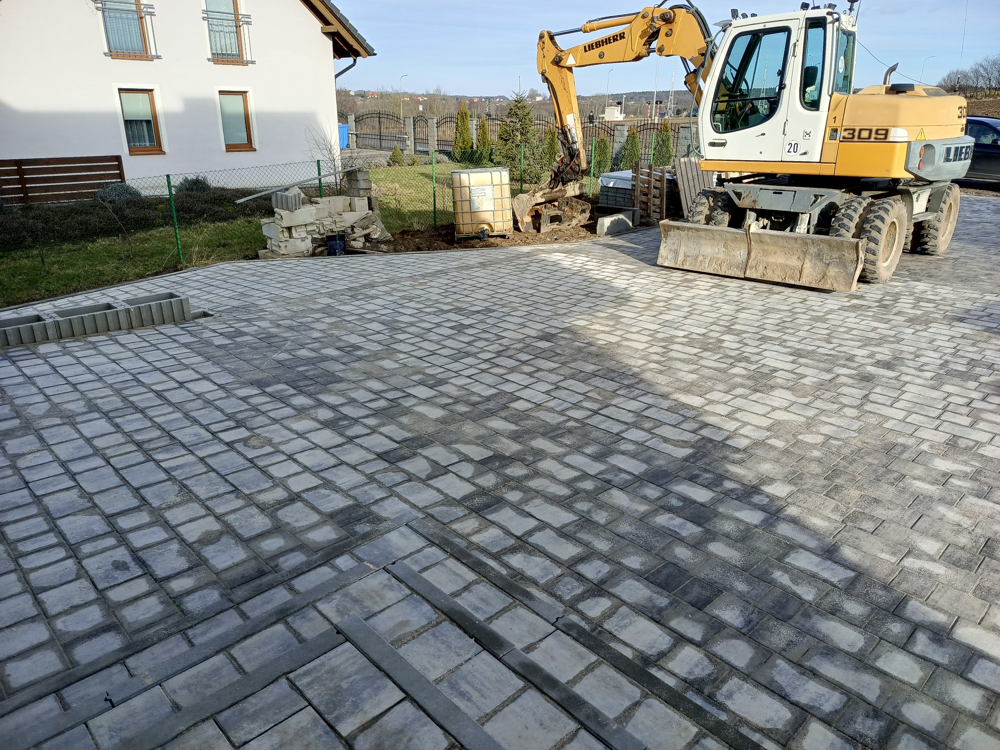 Ułożona szara kostka brukowa na podjeździe przed domem, z żółtą koparką Liebherr w tle, przygotowującą teren pod dalsze prace.