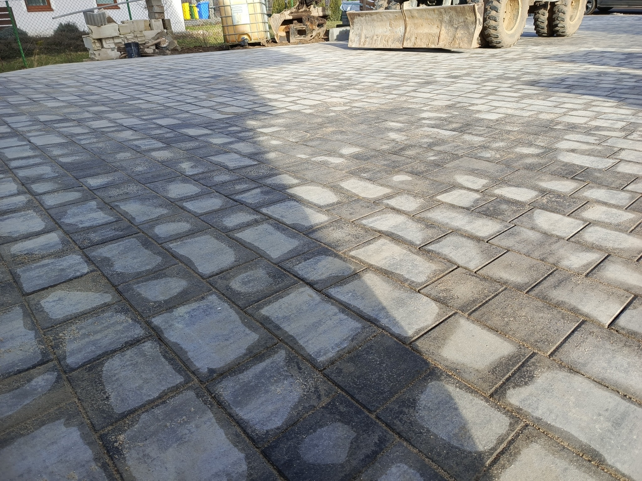 Ułożona, szara kostka brukowa w regularny wzór, z widocznymi śladami wilgoci i piasku w szczelinach, na tle częściowo widocznej maszyny budowlanej i zbiornika.