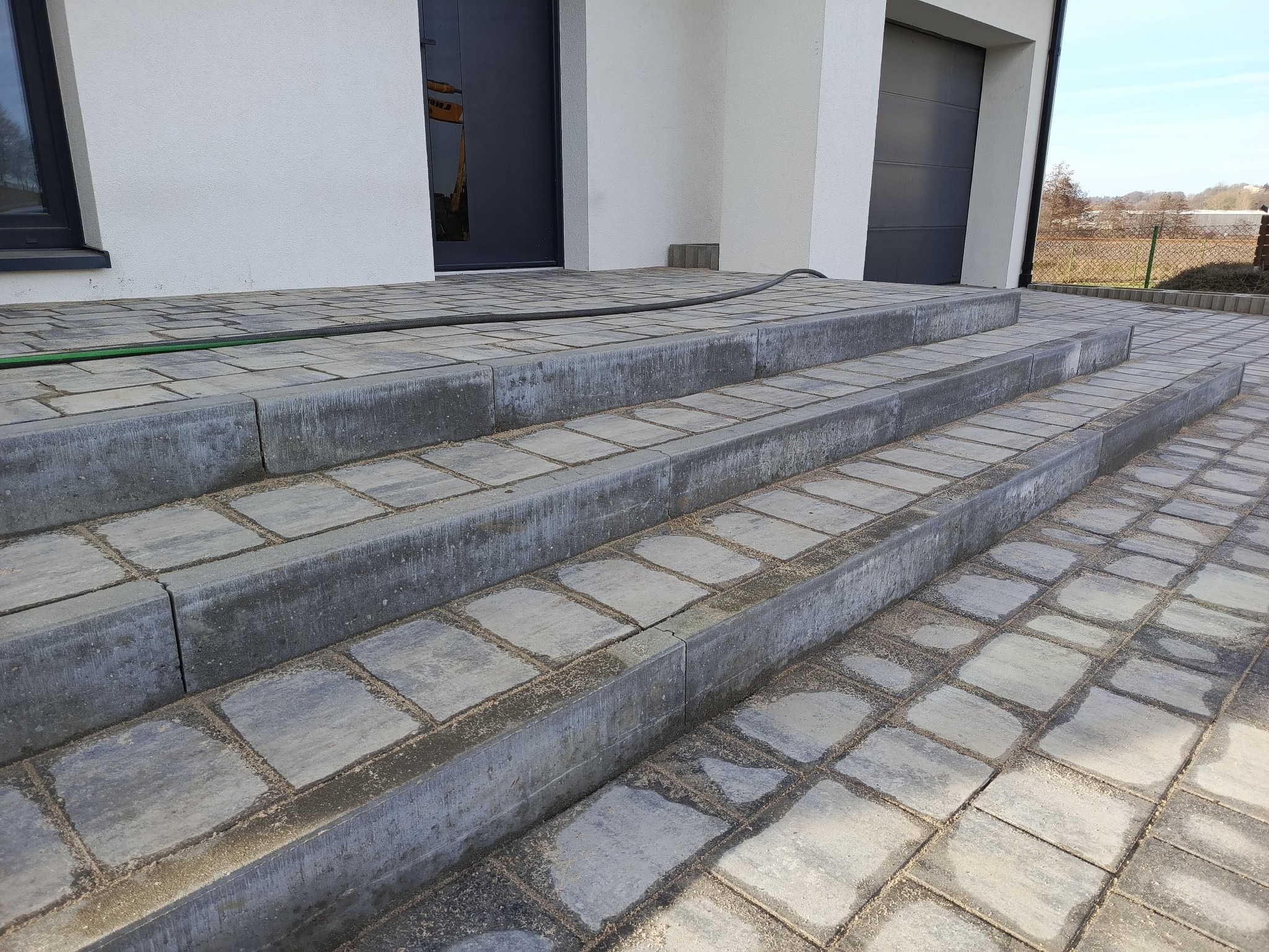 Schody wejściowe do domu wyłożone szarą kostką brukową, z widocznym wężem ogrodowym na górnym stopniu.